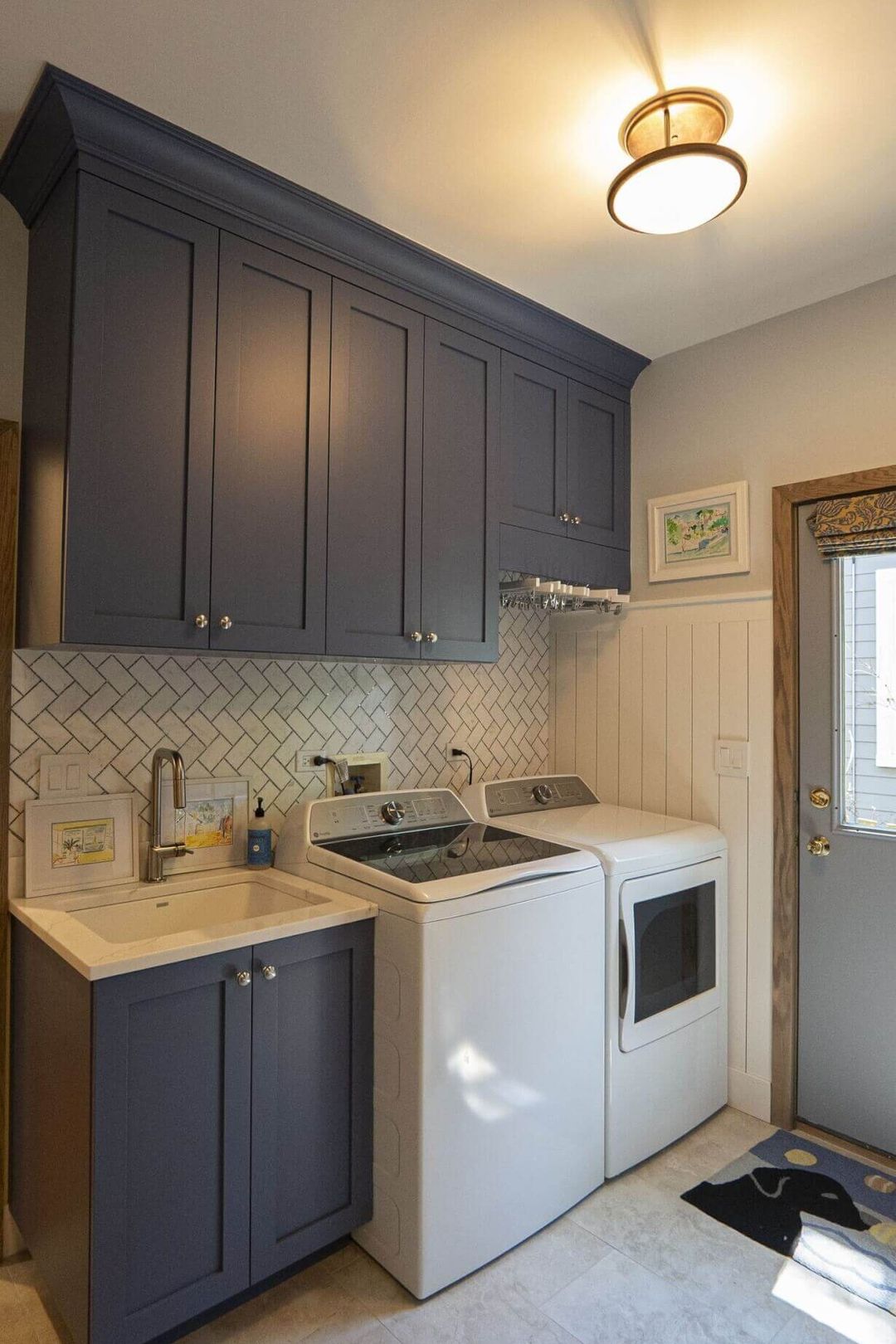 Laundry room with navy shaker cabinets, herringbone tile backsplash, utility sink, shiplap wainscoting, and top loader washer dryer - Kelly Thayer Design