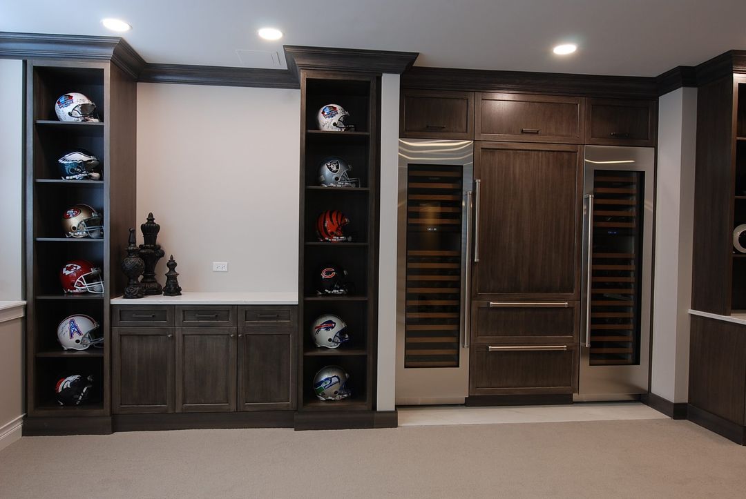 Winnetka lower-level man cave with dark custom built-in shelving, football helmet display, paneled Sub-Zero refrigerator, and wine cooler - Kelly Thayer Design