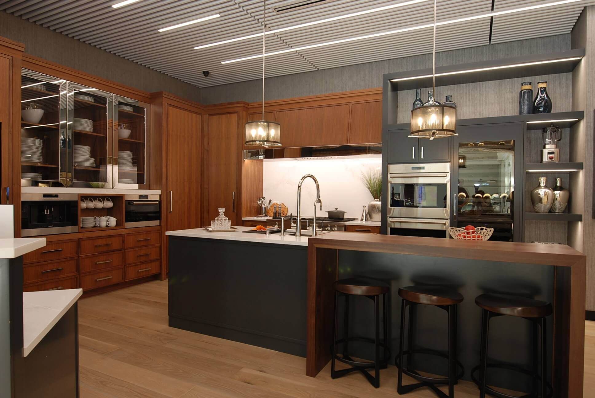 Modern kitchen showroom with wooden cabinets, dark island, and three bar stools under pendant lights.