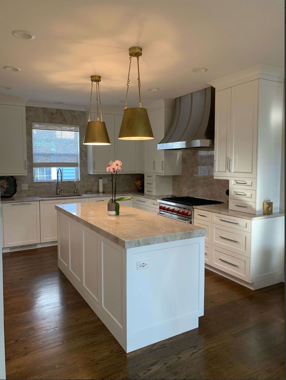White kitchen with a marble-topped island, brass pendant lights, white cabinets, and dark hardwood floors; orchid on the island and a stainless range in the background.